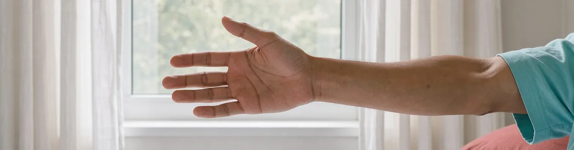An adult's relaxed forearm and open hand extend in a gentle waking stretch beside a bright bedroom window