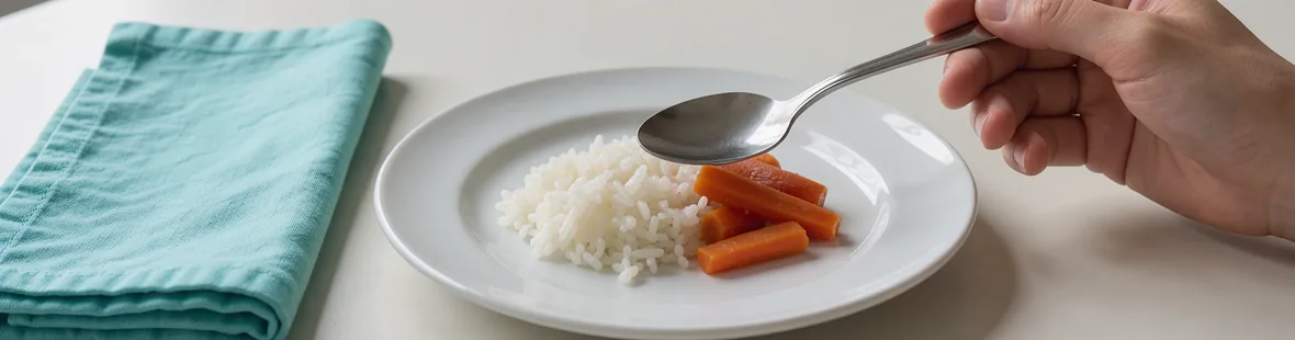 A hand holds a spoon above a small plate of soft rice and cooked carrots on a home table