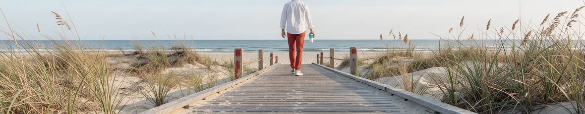 An adult walks a long Long Island beach boardwalk in early morning light, coffee in hand.