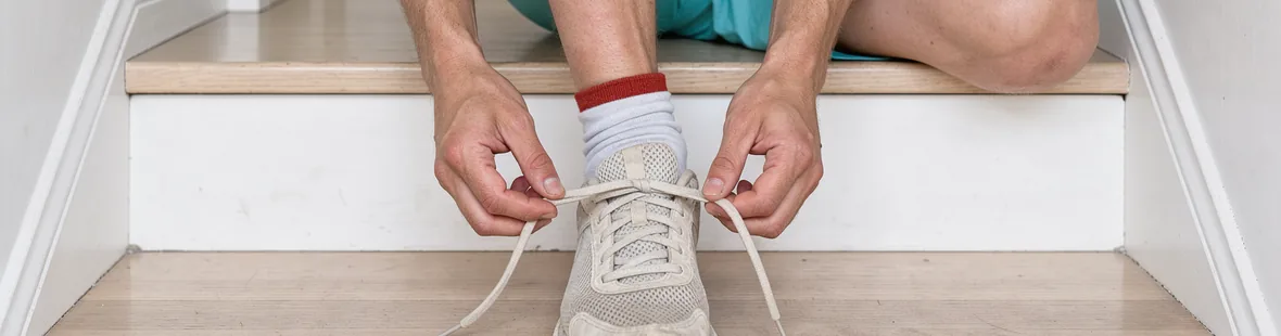 A seated runner loosens an unmarked running shoe on a low step at home, with relaxed hands and dusty socks